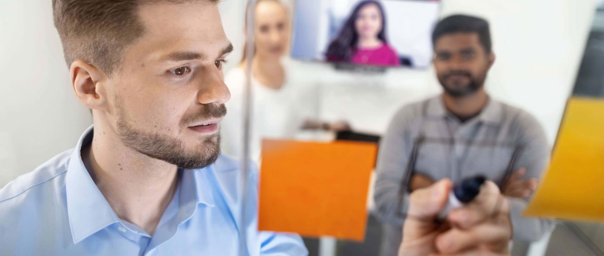 A person discussing notes on a glass pane with colleagues; other people blurry in the background and a person joining digitally